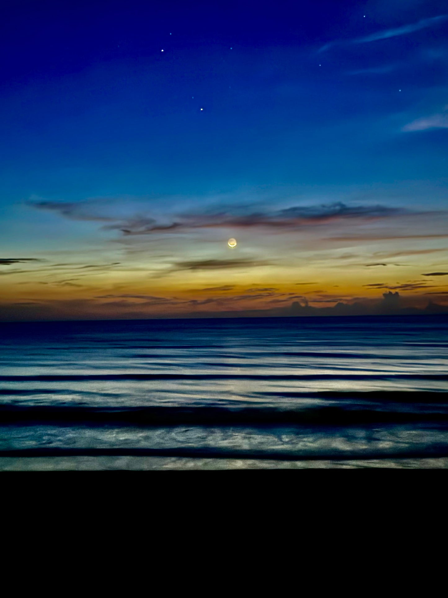 A serene seascape at dusk or dawn with dark blue water in the foreground, a glowing yellow moon near the horizon, and a deep blue sky with faint clouds and stars above.