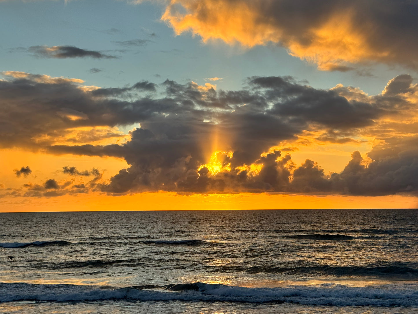 A dramatic sunset over the ocean with golden light breaking through clouds, dark stormy clouds across the sky, and waves rolling onto the shore.