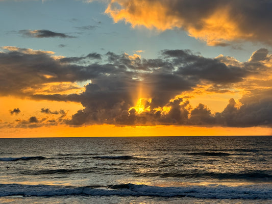 A dramatic sunset over the ocean with golden light breaking through clouds, dark stormy clouds across the sky, and waves rolling onto the shore.