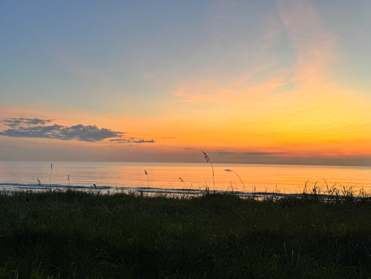 A scenic sunset over a calm body of water with a dark grassy foreground, showing a gradient sky from blue to orange and yellow.