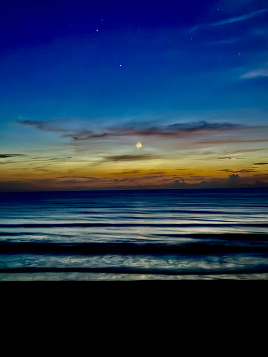 A serene seascape at dusk or dawn with dark blue water in the foreground, a glowing yellow moon near the horizon, and a deep blue sky with faint clouds and stars above.