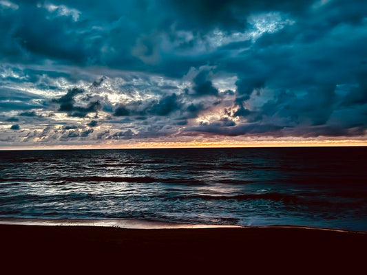 A dramatic seascape at sunset with dark blue and teal storm clouds filling the sky, glowing orange light along the horizon, rippling water, and a dark sandy shore in the foreground.