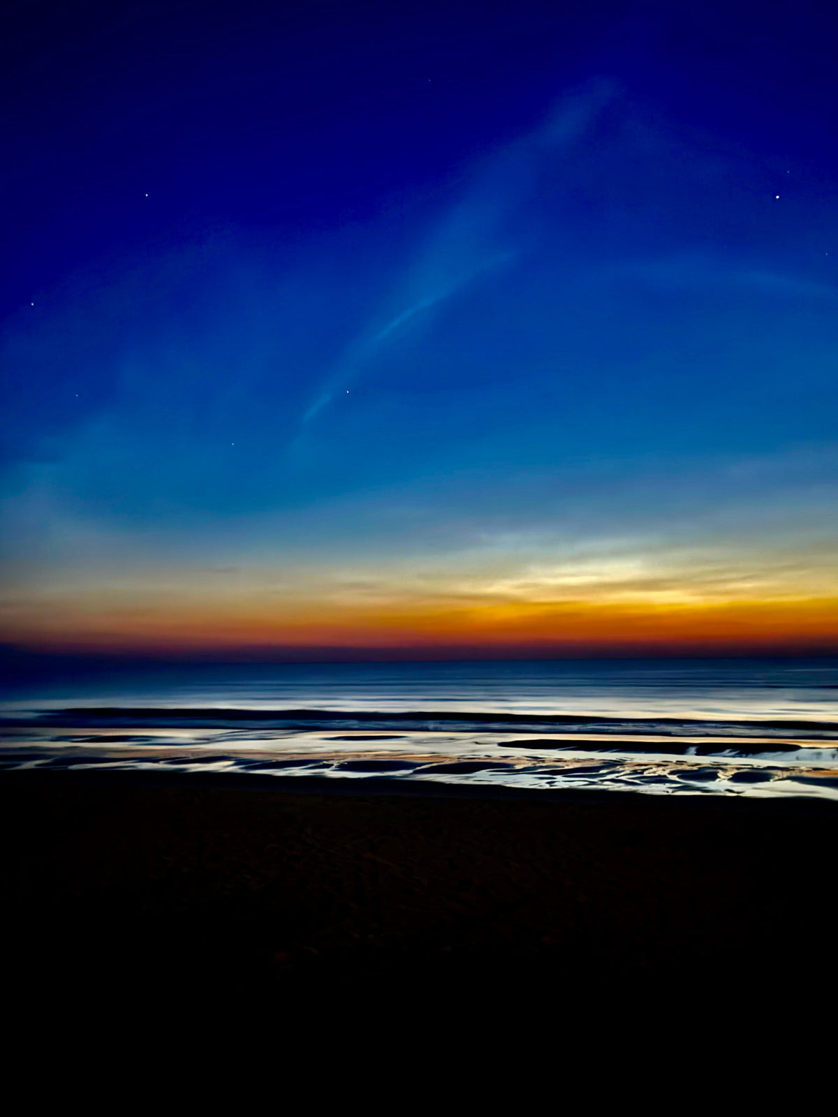 A scenic landscape photograph of a beach at twilight, featuring a dark silhouetted shoreline and tidal flats in the foreground, with a gradient sky transitioning from deep blue to orange and yellow near the horizon.